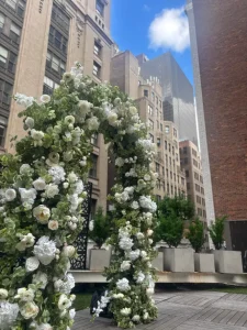 An intimate rooftop ceremony in progress with guests seated in white chairs facing a floral arch and the officiant.